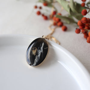 handmade small gold oval pendant sits against a white background with bright red berries in background