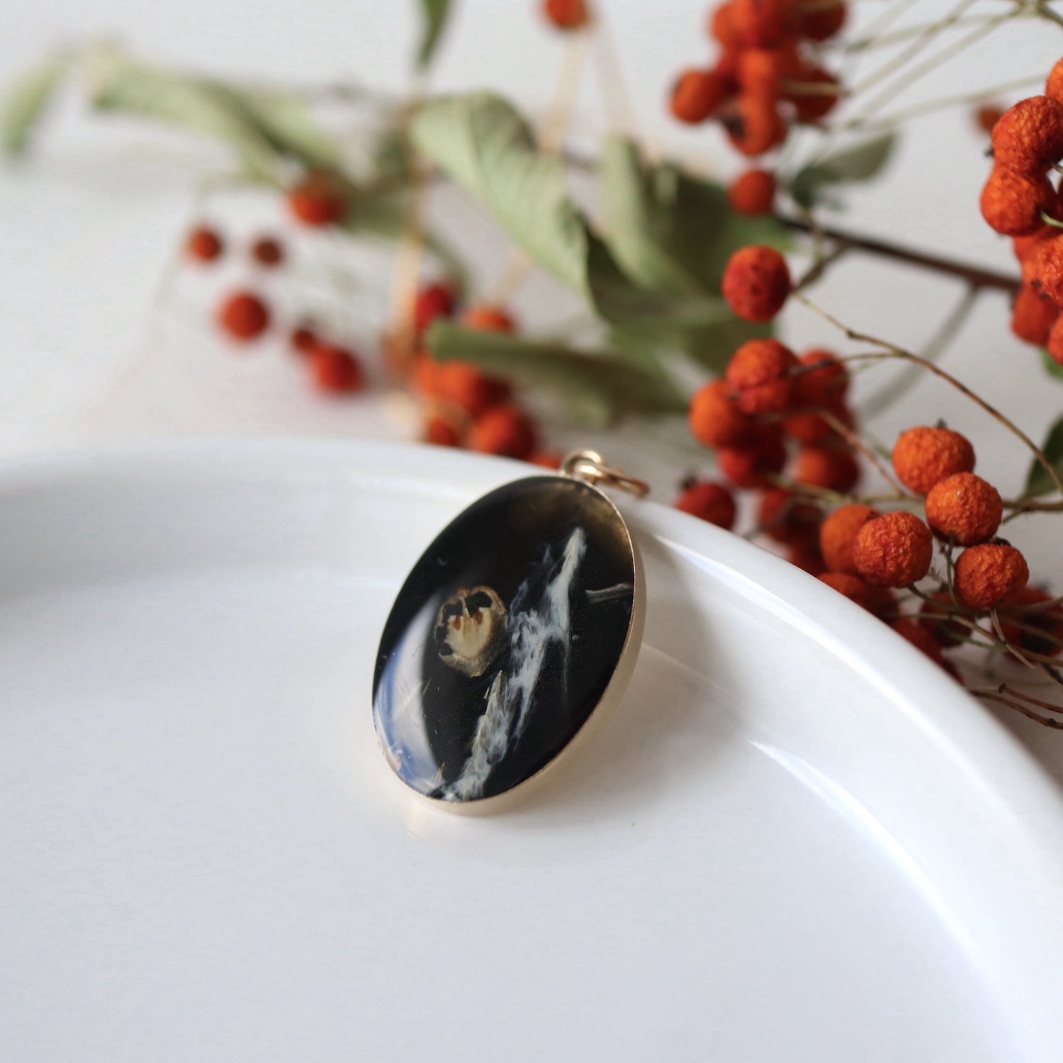 handmade small gold oval pendant sits against a white background with bright red berries in background