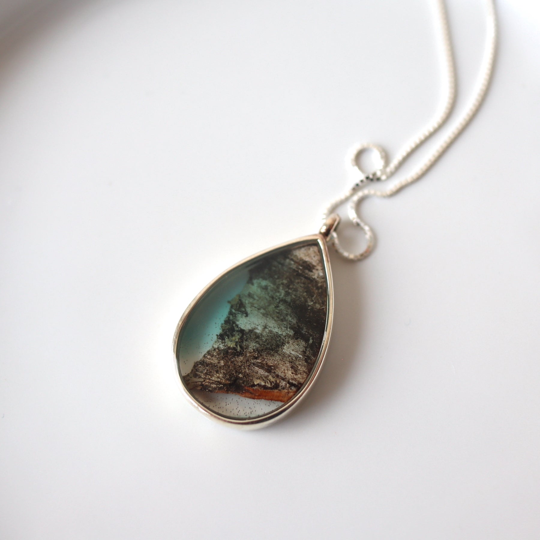 sterling silver teardrop frame encases a fragment of birch bark in softly tinted resin. The necklace sits on a white ceramic background. 